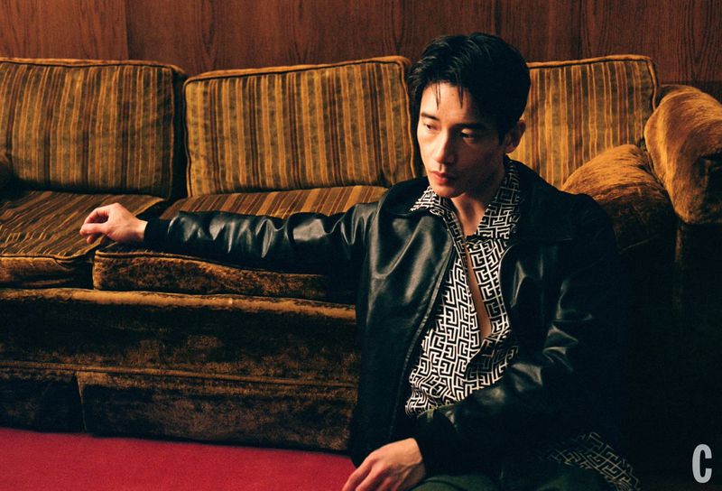 A stylish Asian man poses in a black leather jacket and patterned shirt, seated on a red floor against a vintage couch and wood panel wall.