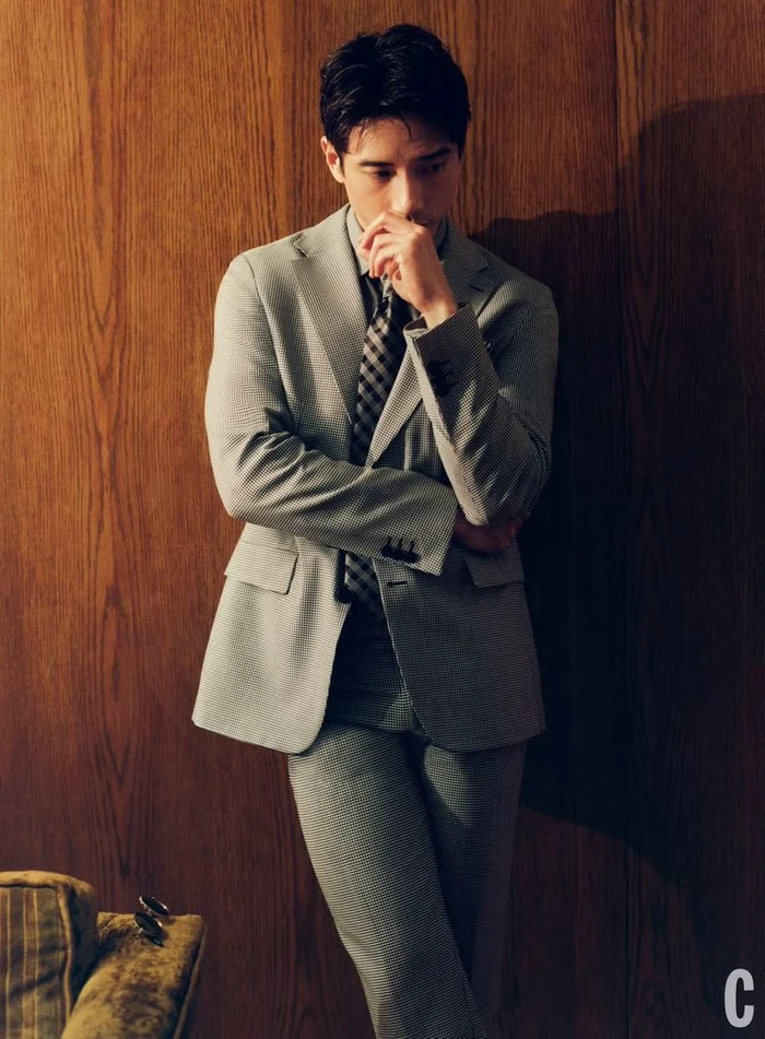 Manny Jacinto leaning against a wood paneled wall wearing a grey suit and patterned tie for Cosmopolitan U.S.