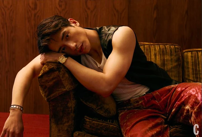 Manny Jacinto leaning on a velvet sofa wearing a leather vest and patterned red pants for Cosmopolitan U.S.