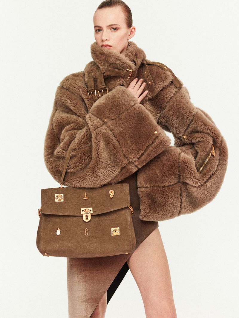 Fashionable woman poses in a studio wearing a fur coat and holding a stylish brown leather handbag with gold hardware.