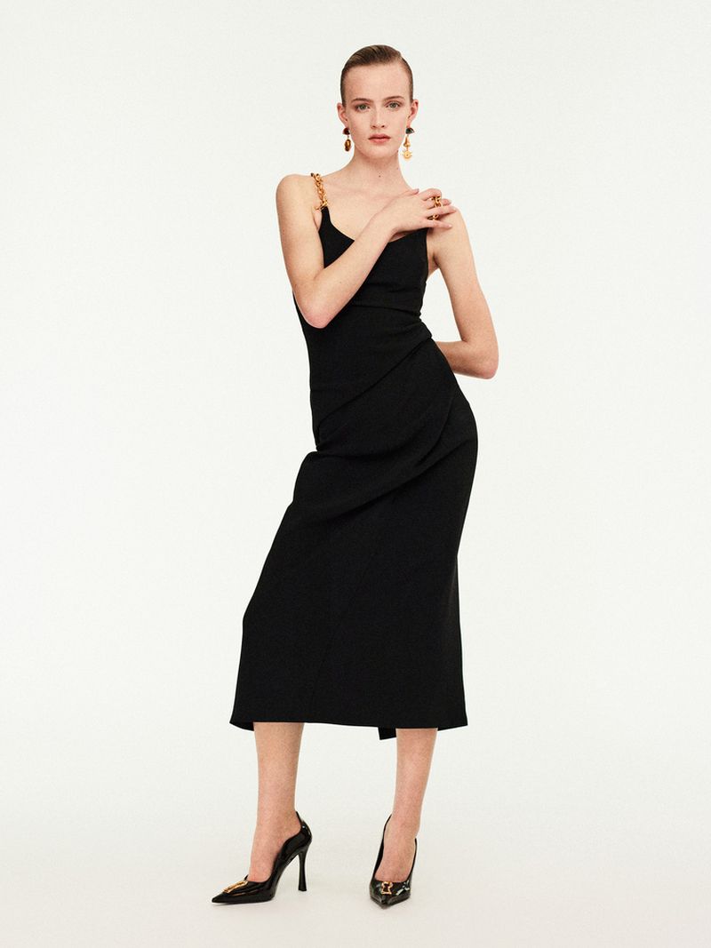 Elegant woman in a black dress and heels poses against a white backdrop, showcasing fashion and style with chic accessories and poise.