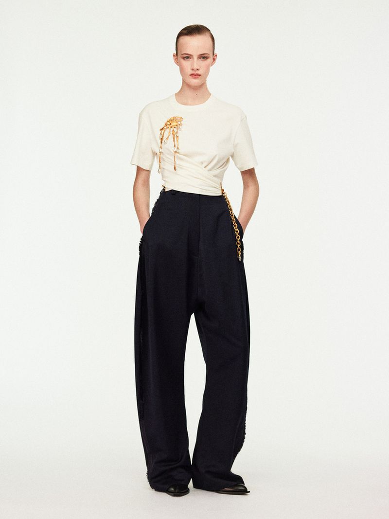 Fashion shot of a slim adult woman in wide-leg black trousers and a cream-colored t-shirt with a gold chain detail, posing indoors.