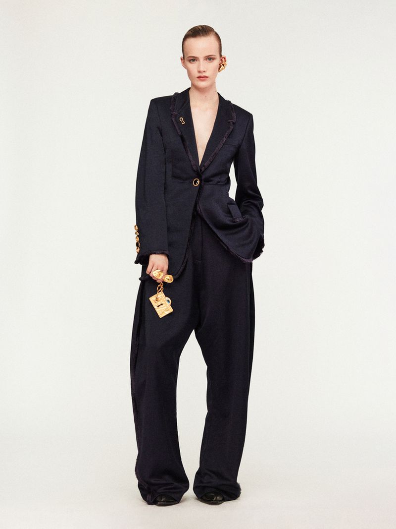 Fashion editorial shot of a woman posing in an oversized dark blue suit with bold gold earrings and key chain against a white backdrop.