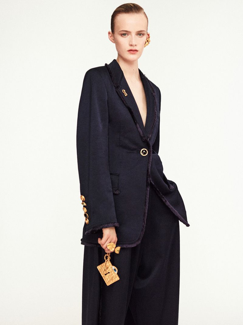 Fashion model poses in a navy blazer and wide-leg pants with gold accessories against a white background in a studio shot.