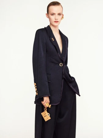Fashion model poses in a navy blazer and wide-leg pants with gold accessories against a white background in a studio shot.