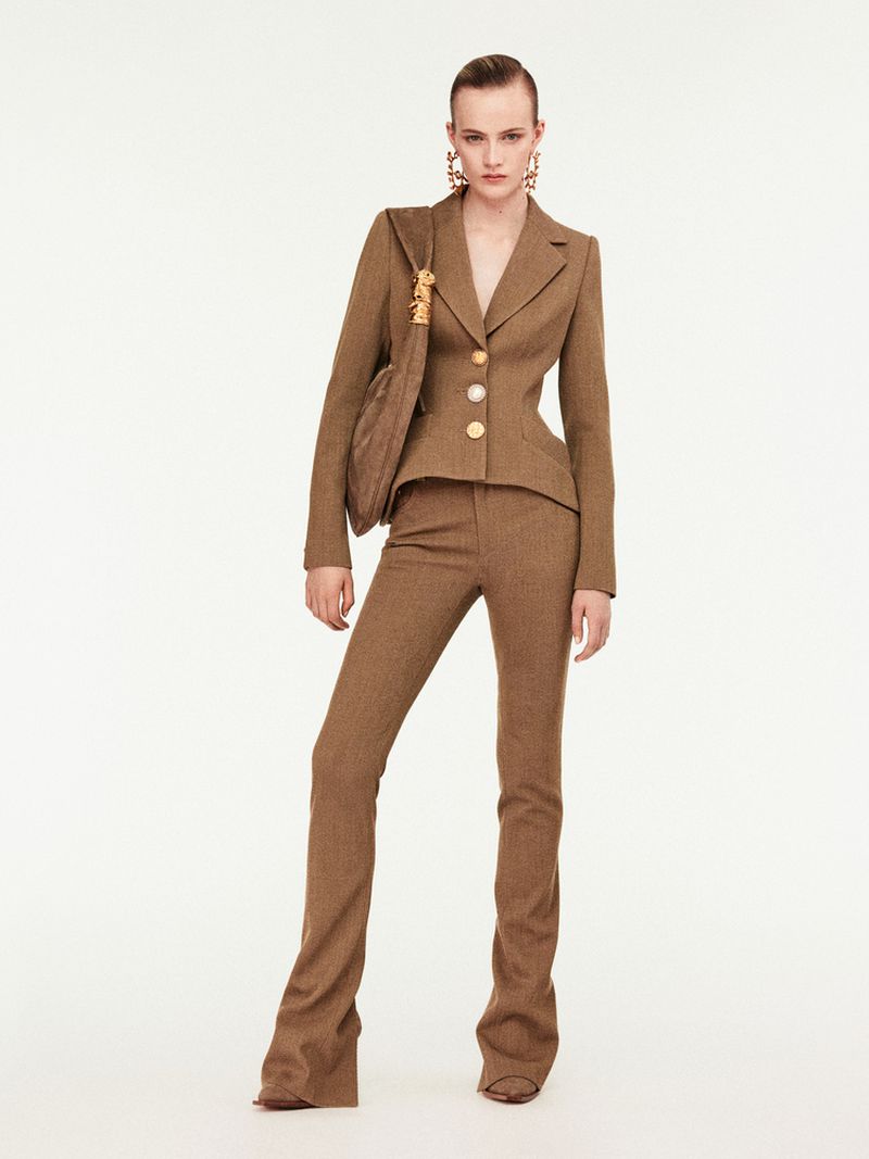 Fashion model in brown tailored suit and large earrings, carrying a brown fabric handbag, posing against a white backdrop in a studio.