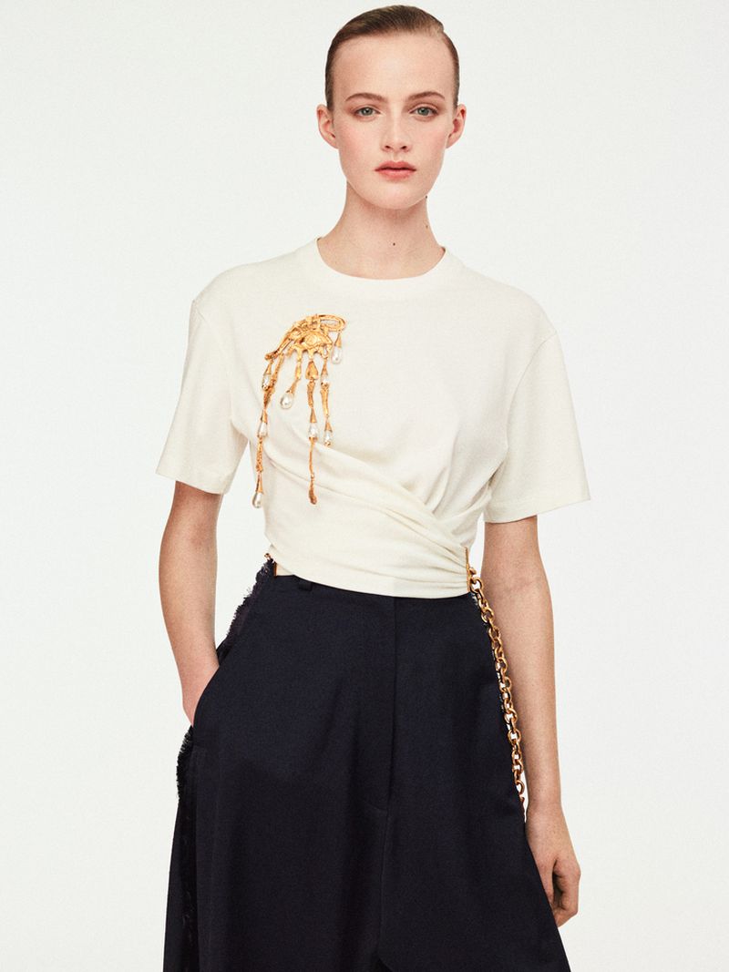 Fashion shot of an adult woman in a white t-shirt with a gold pendant and navy wide-leg pants against a white backdrop.
