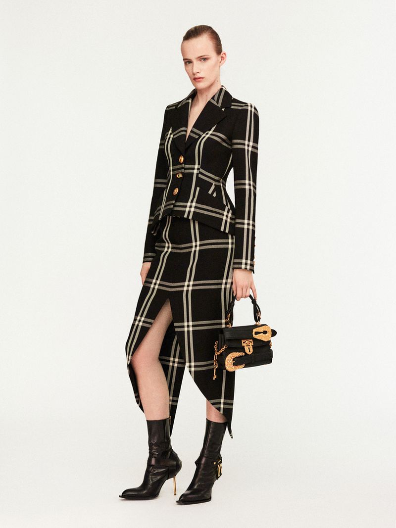 Fashionable woman in a black-and-white plaid blazer and skirt, holding a purse and wearing heeled boots, posing in a photo studio.