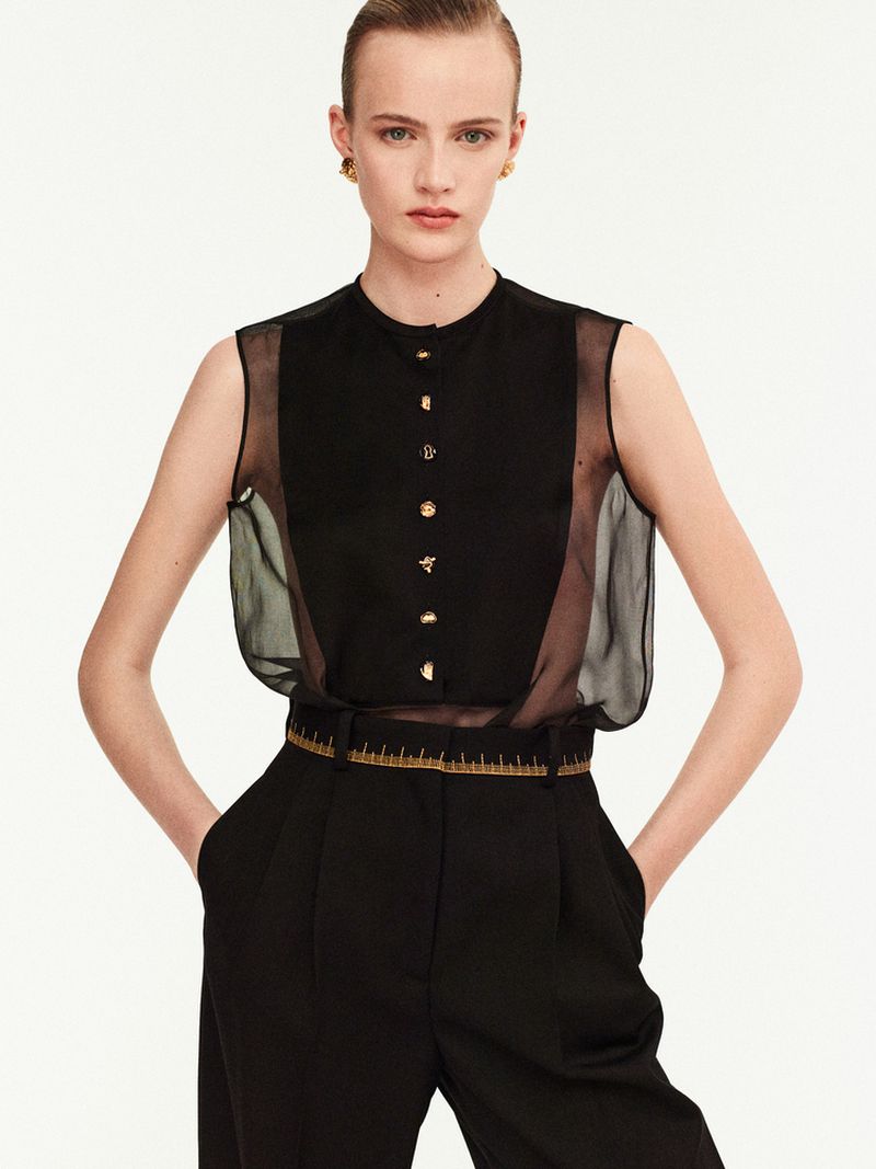 Fashion portrait of a fair-skinned woman with blonde hair wearing a black sleeveless top and trousers against a white backdrop.