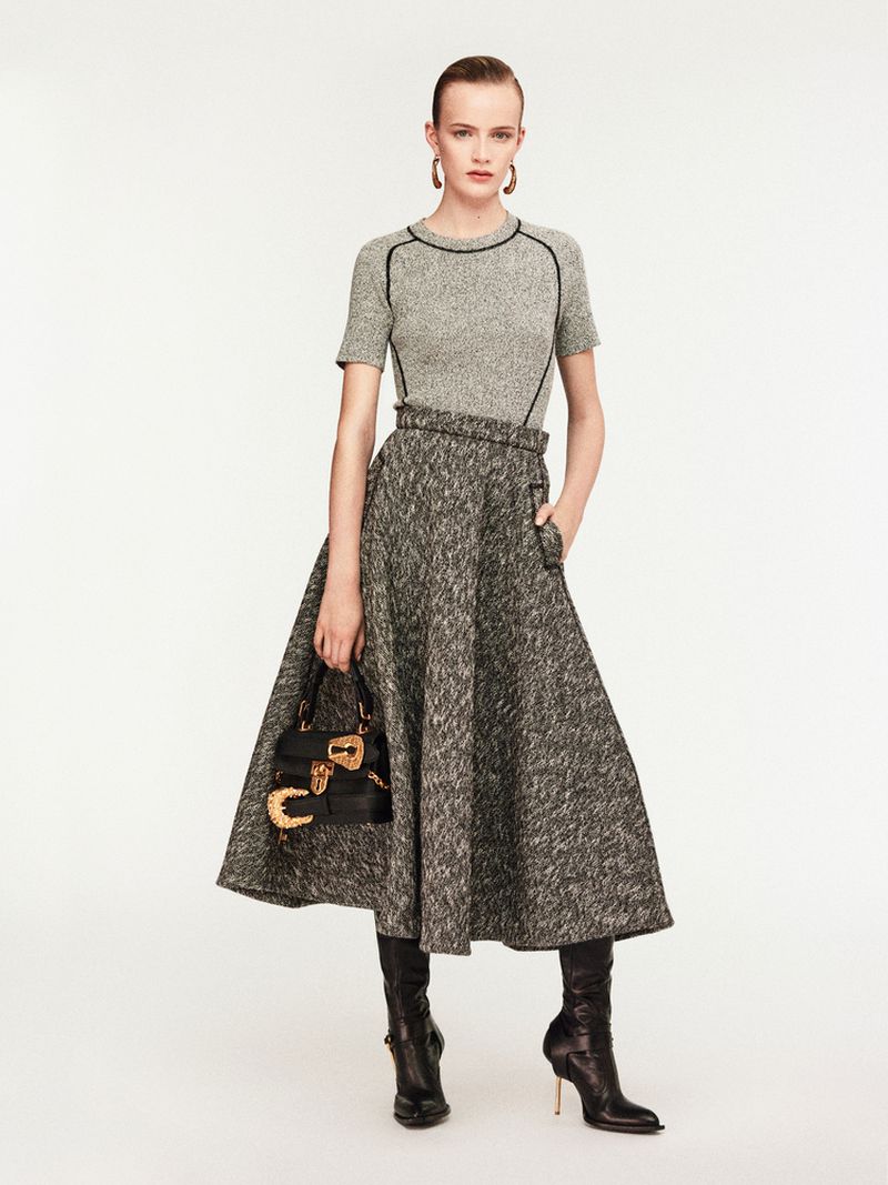 Full-length studio shot of a woman in a gray tweed midi skirt and matching top, holding a black leather purse and wearing heeled boots.