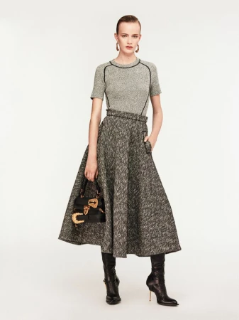 Full-length studio shot of a woman in a gray tweed midi skirt and matching top, holding a black leather purse and wearing heeled boots.