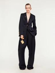 Model wearing a dark tailored suit and gold statement accessories against a plain white studio backdrop.