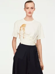 Model wearing a white t-shirt with a gold brooch and dark trousers against a plain white background for Schiaparelli F/W25 Lookbook