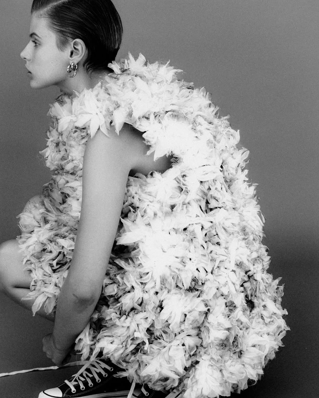 Black and white portrait of a young woman in a feather-like dress, crouching with sneakers, against a neutral gray backdrop.