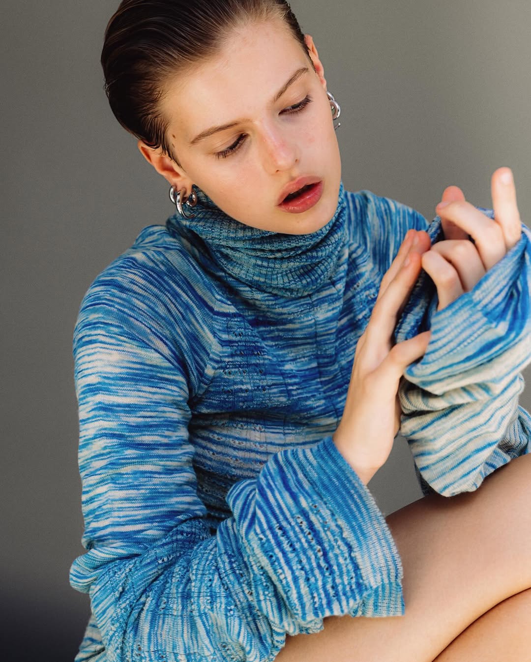 Young woman with wet-look hair, wearing a blue patterned turtleneck sweater, poses indoors against a neutral background in a close-up shot.