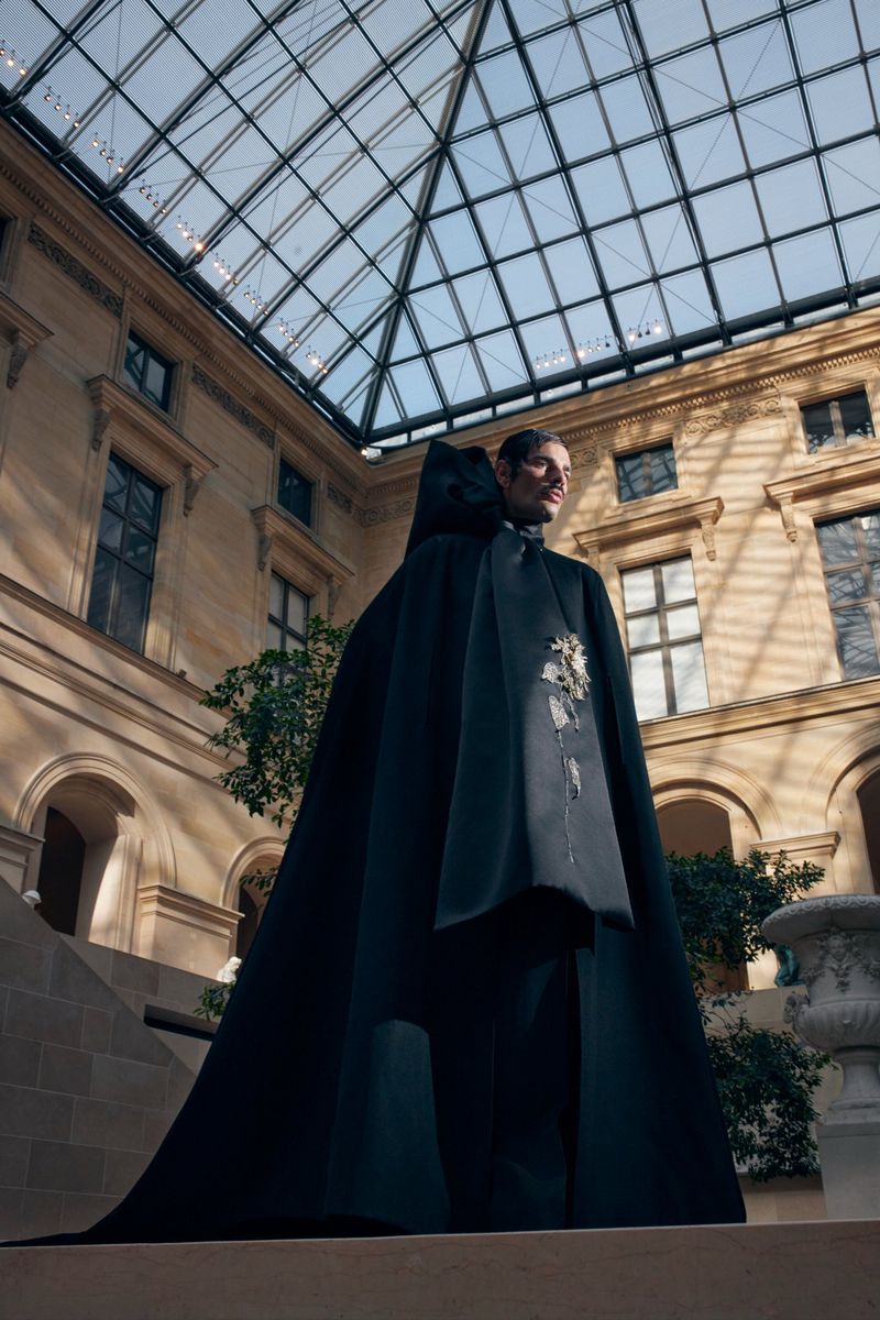 Fashion shot of a man in a formal cape standing indoors, featuring architecture with a glass ceiling and trees in the background.