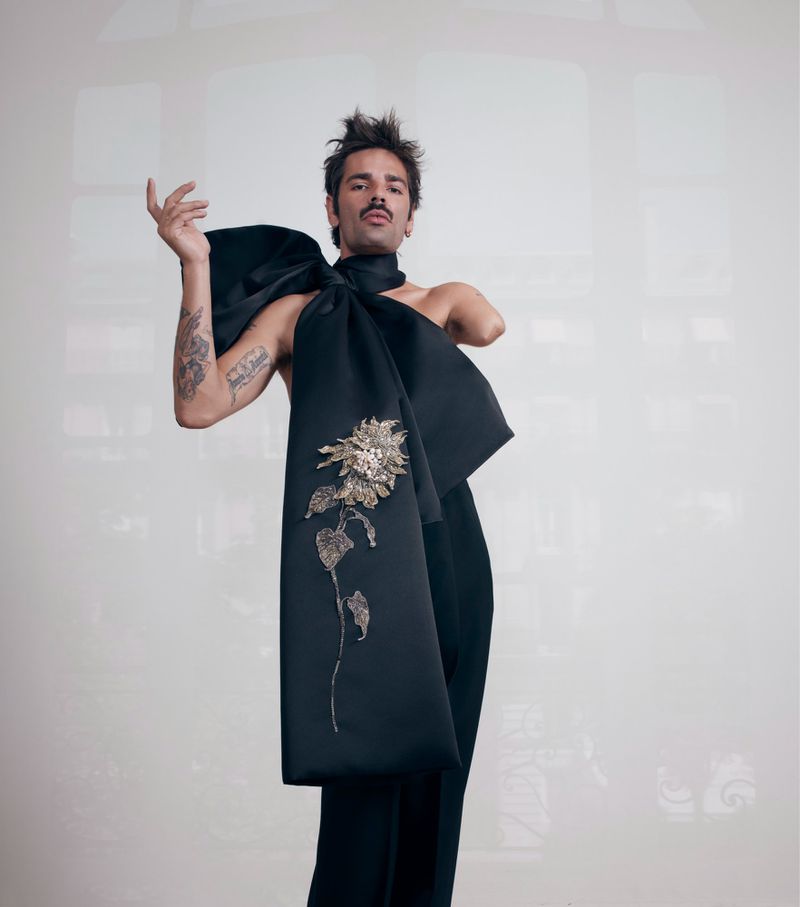 Adult male with mustache posing in studio wearing black pants and sculptural one-shoulder top with floral embroidery on a solid background.
