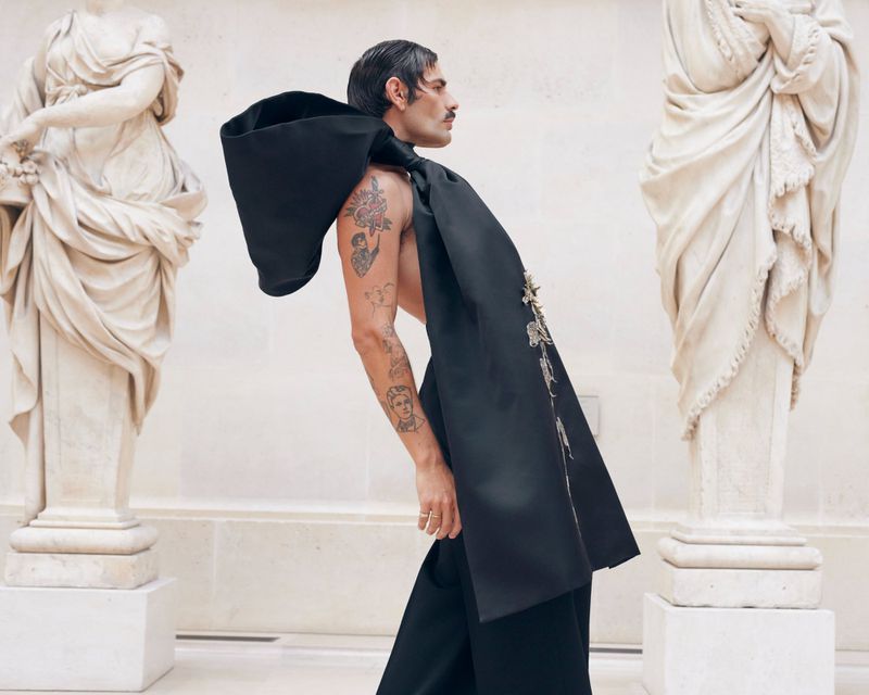 A man with tattoos and a mustache strikes a pose in an oversized black hooded garment. Statues are on pedestals in the background.