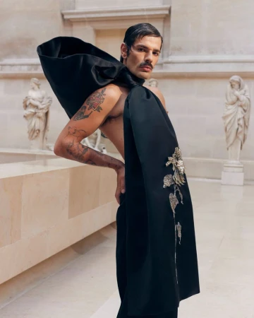Man posing in high fashion outfit with floral embellishments, set against a backdrop of classical sculptures and architectural details.