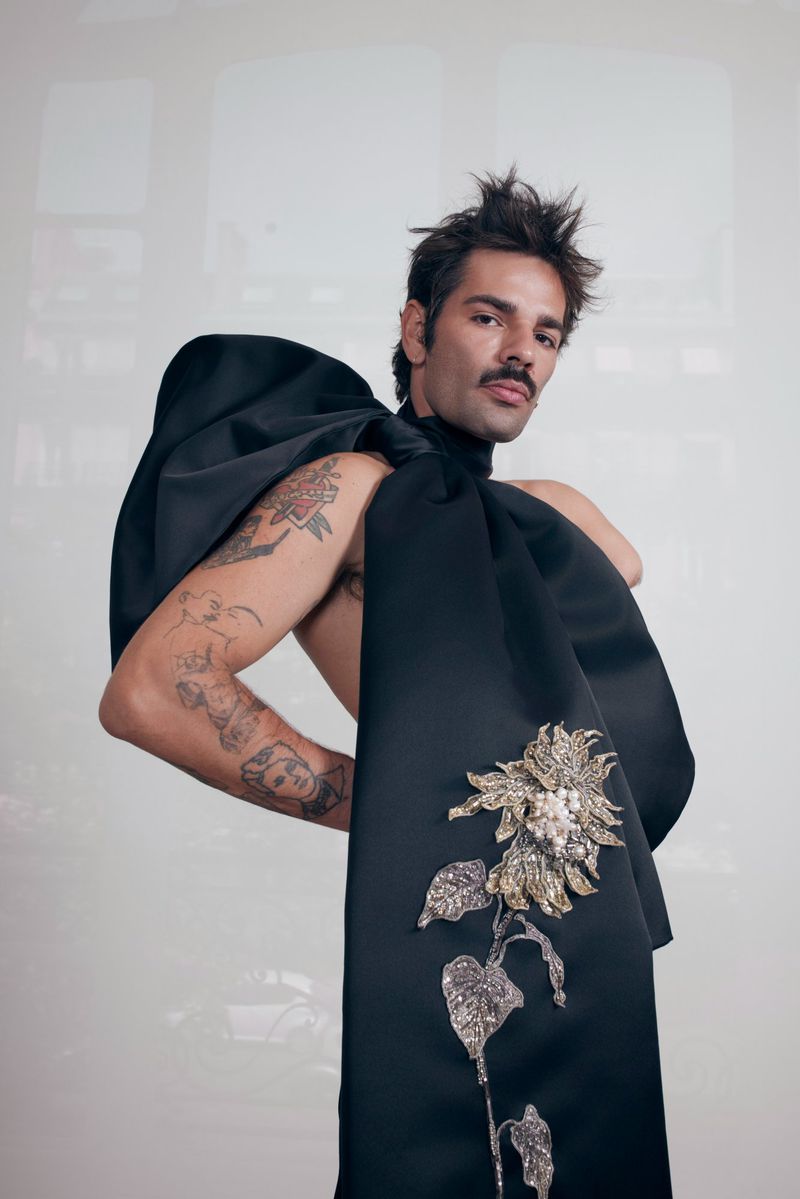 Man poses in black formalwear with a large bow-like sash adorned with a sunflower brooch, against a bright studio background.