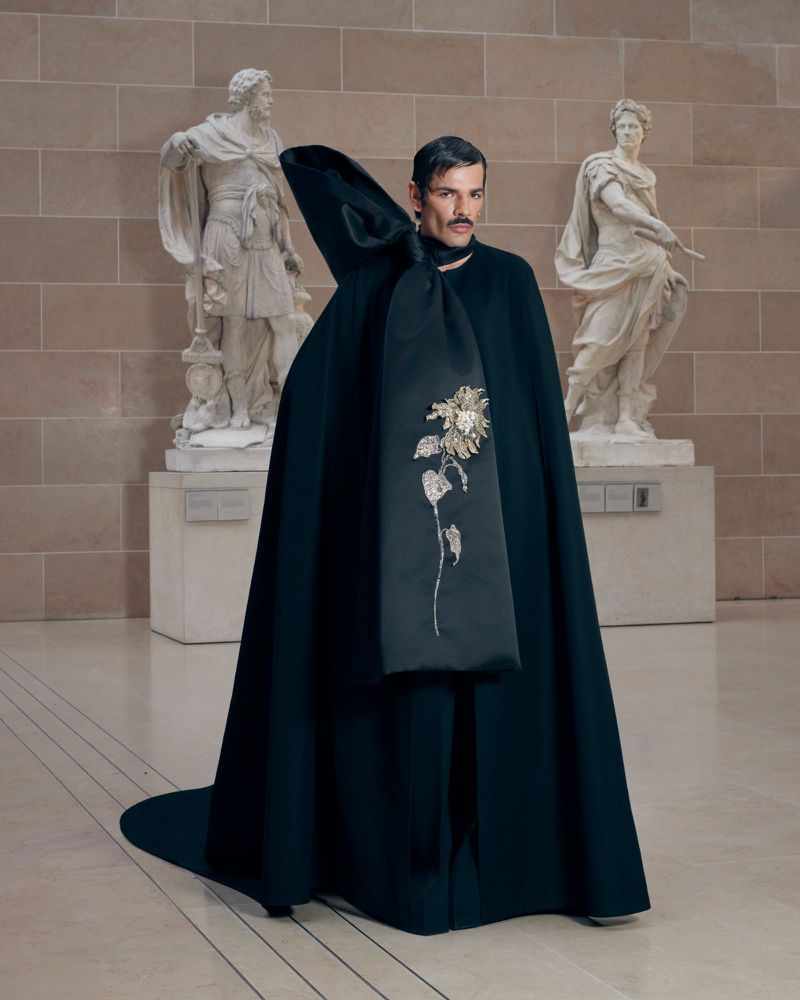 Fashionable adult male poses indoors wearing long, dark cape with floral embellishments, with statues in background in museum setting.
