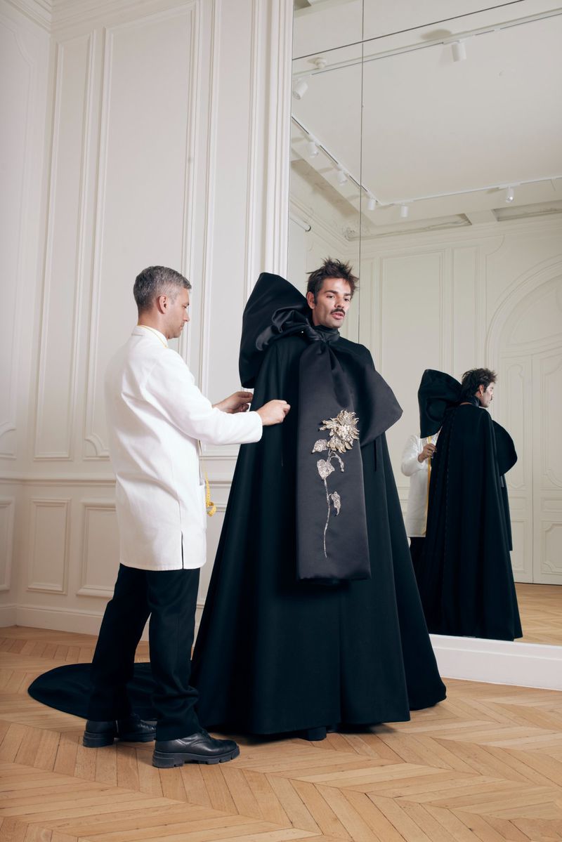 A designer fits a large black cape with an oversized bow on a man in a studio with a herringbone floor and mirror backdrop.