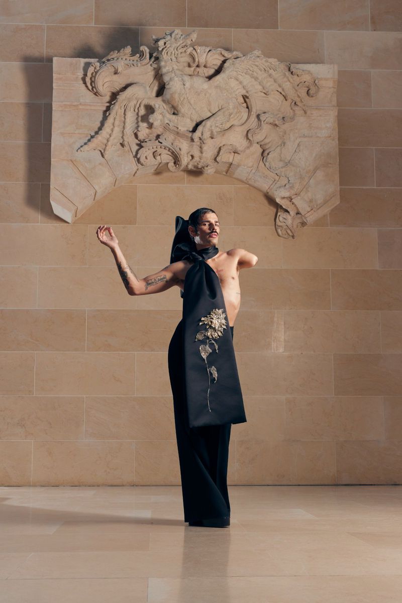 Person in avant-garde fashion poses in interior, wearing black outfit and headpiece with decorative floral design against stone wall.