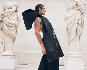 Man wearing a black sculptural outfit with a large back bow standing between classical stone statues