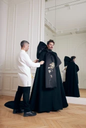 Lucky Love stands in a black embroidered Givenchy haute couture cape while an atelier tailor in a white coat adjusts the fitting.