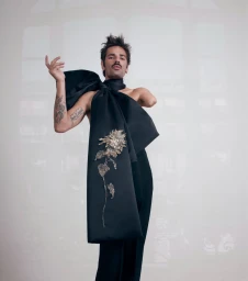 Man with tattoos wearing a black sash and trousers with an embroidered floral embellishment against a bright studio background