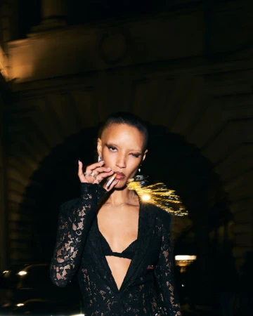 Model Georgia Palmer smoking a cigarette in a black sheer lace blazer at night with warm city lights in the background.