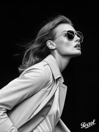 A black and white studio shot of a woman with fair skin wearing sunglasses and a beige trench coat against a solid black background.