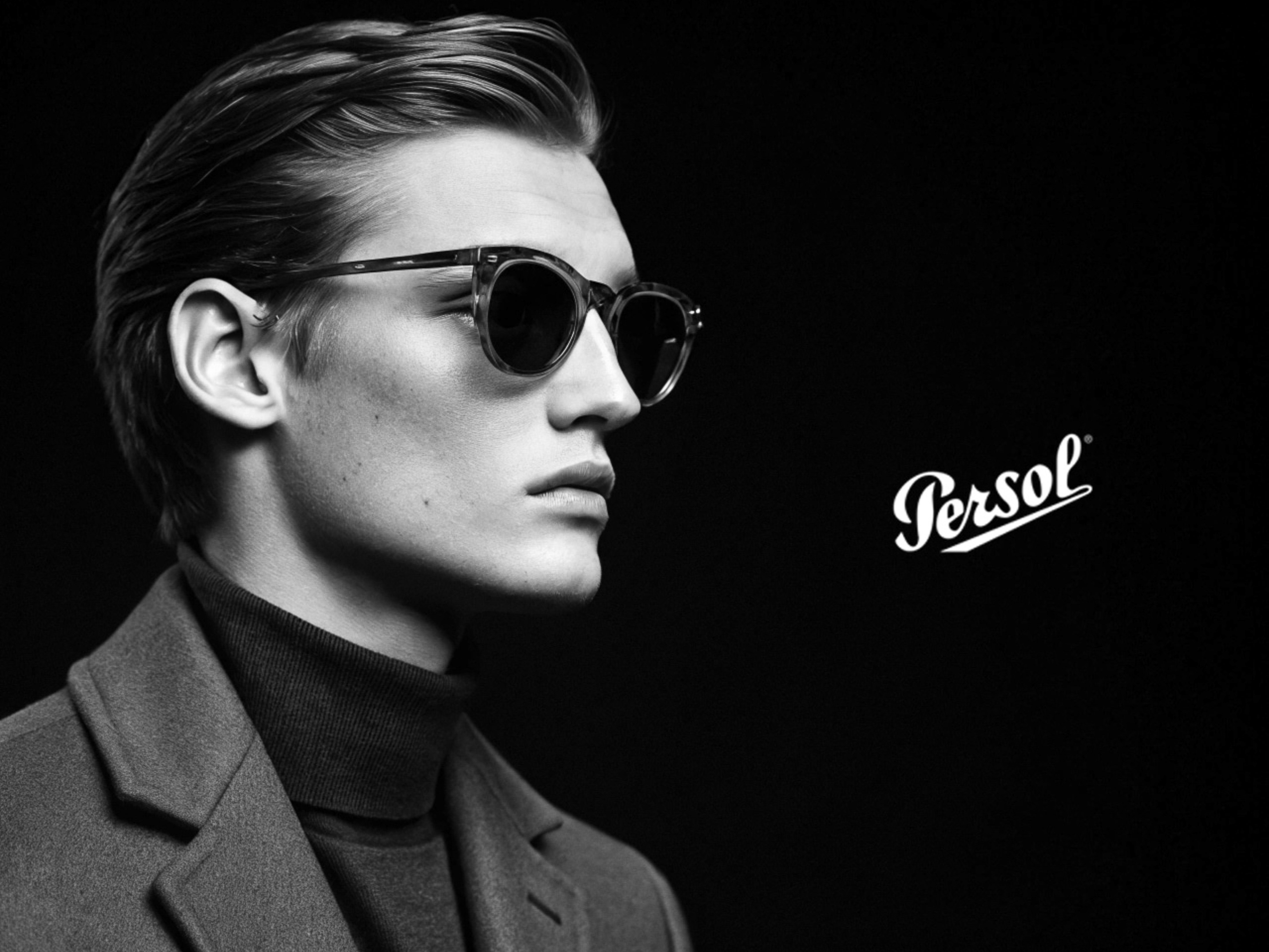 Black and white portrait of an adult male model wearing sunglasses and a coat with a 'Persol' logo in the black background.