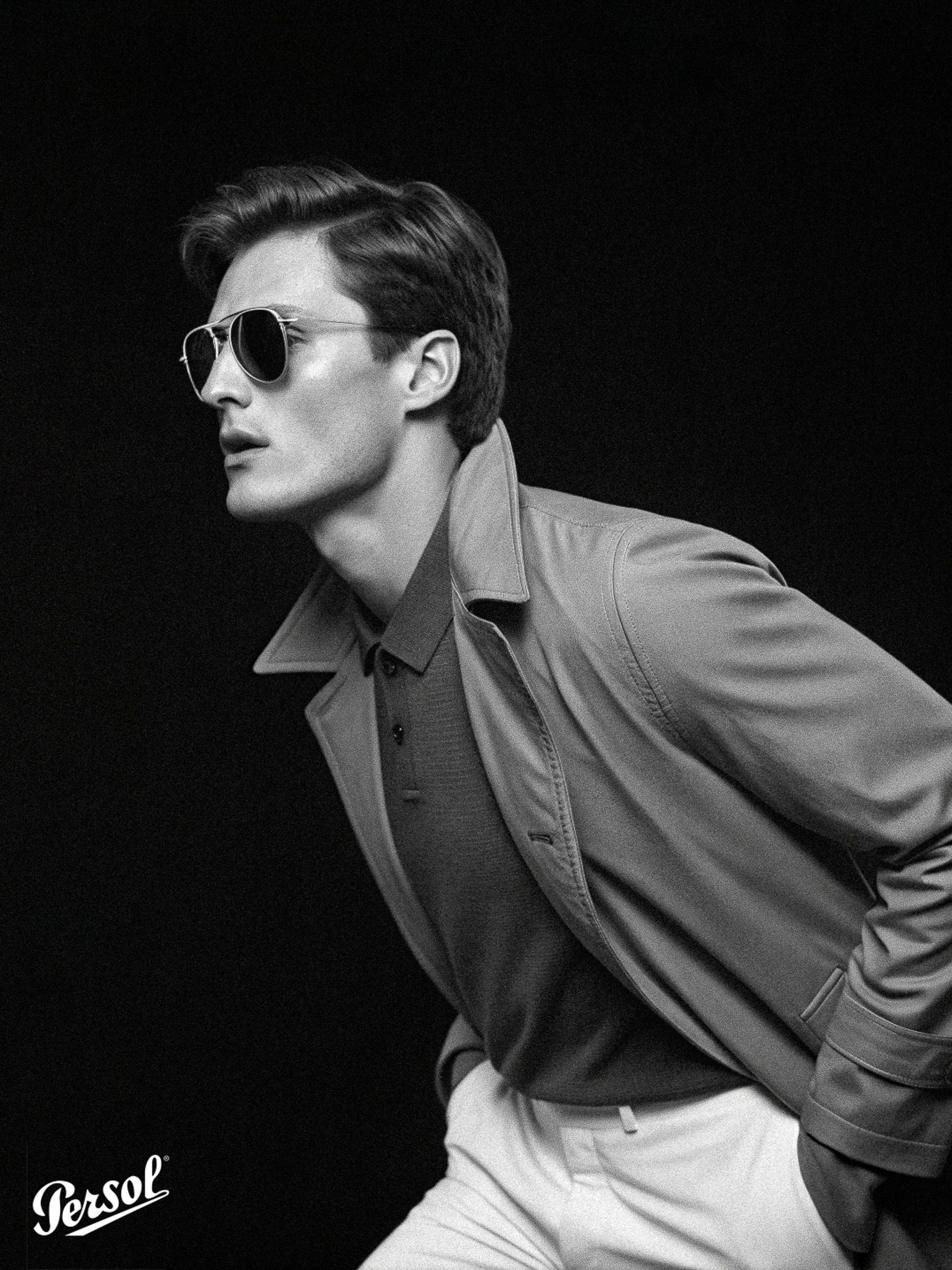 Black and white headshot of a handsome adult male wearing aviator sunglasses, polo shirt, and light trench coat in a studio.