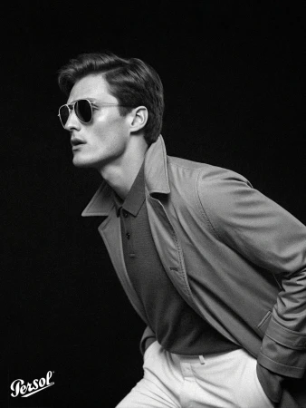 Black and white headshot of a handsome adult male wearing aviator sunglasses, polo shirt, and light trench coat in a studio.