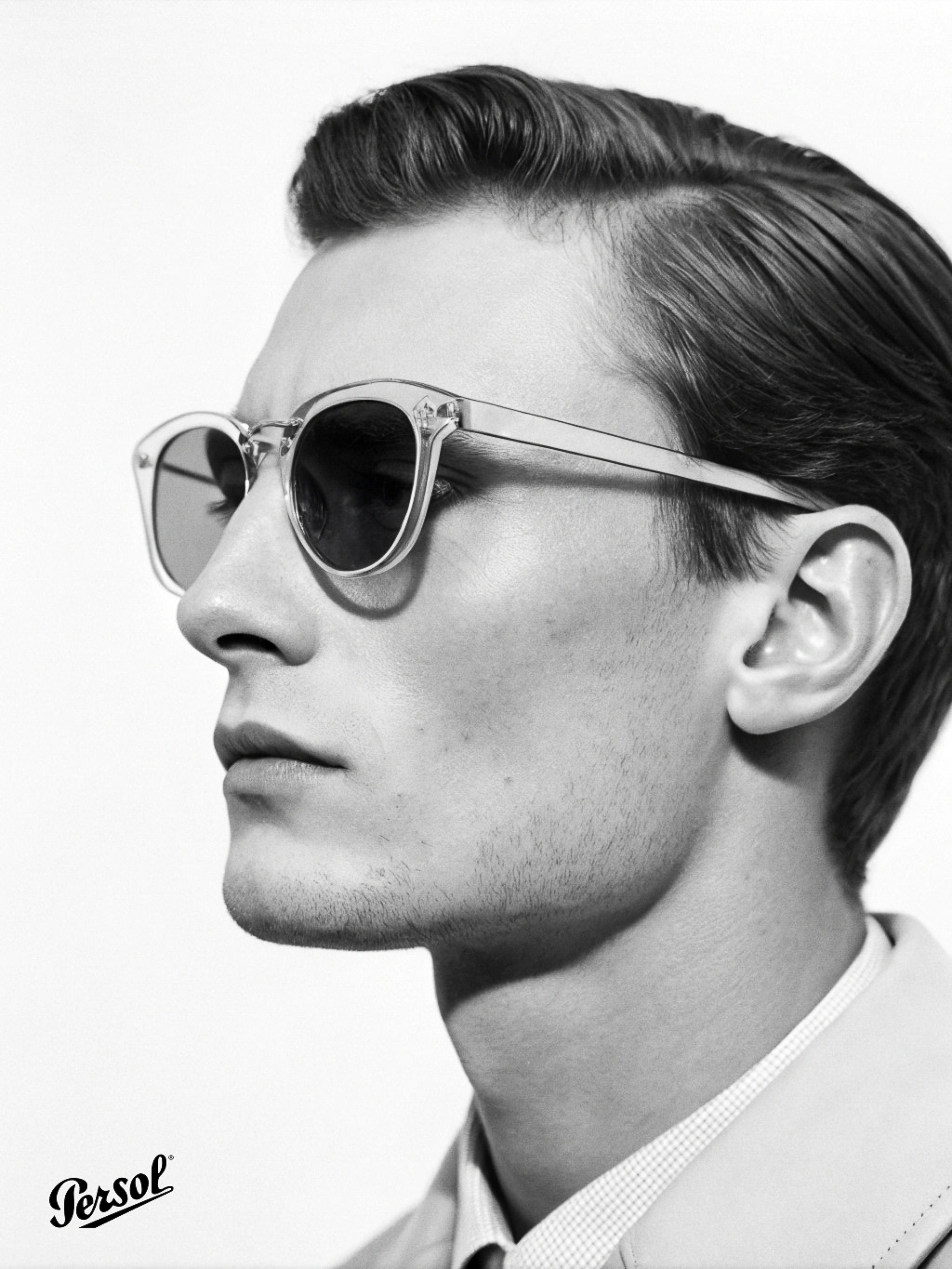 Black and white profile portrait of a man wearing clear acetate sunglasses against a plain white background with a Persol logo.