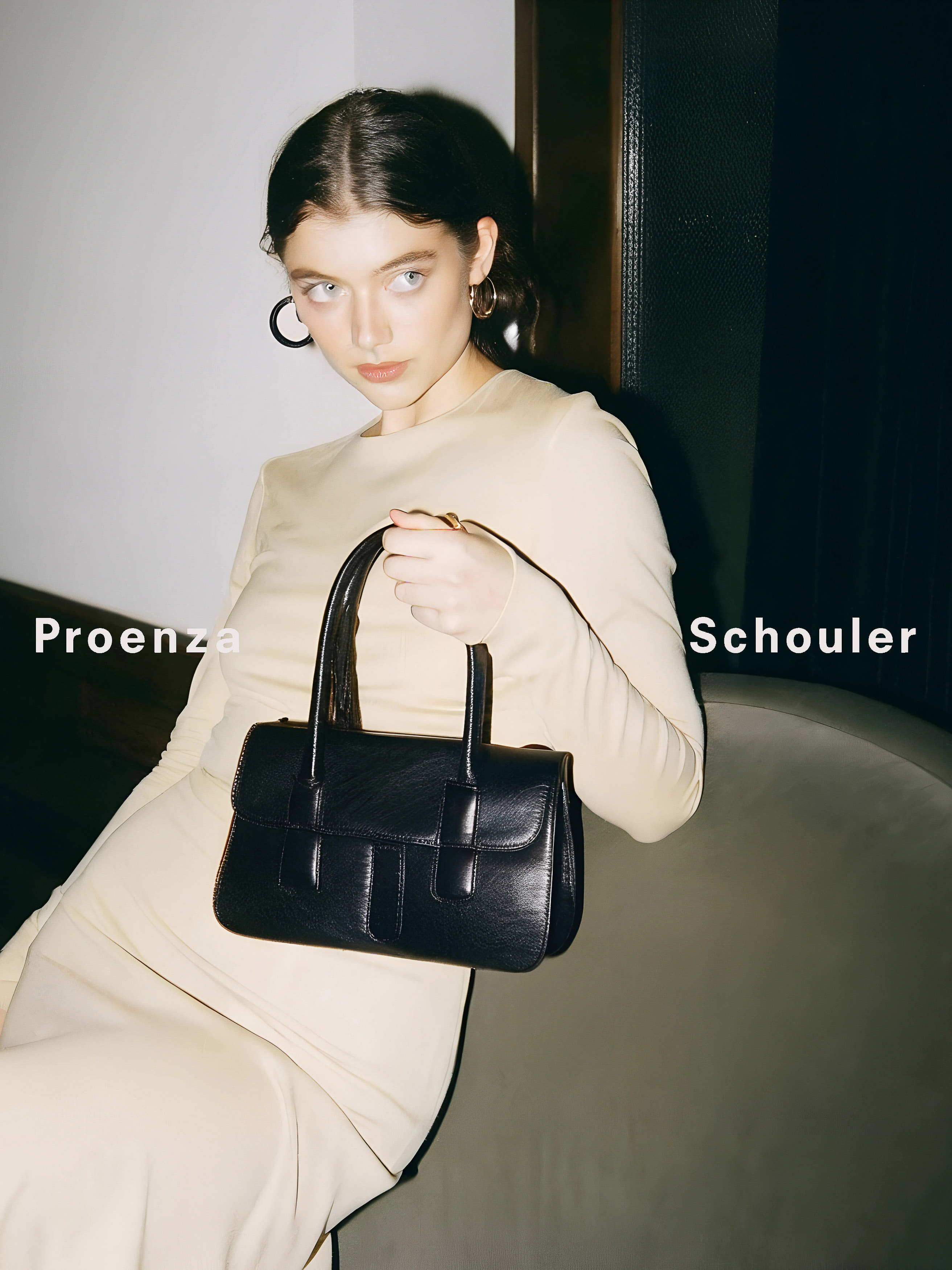 Brunette model holds a black leather handbag in an indoor fashion studio portrait, with visible Proenza Schouler branding on the side.