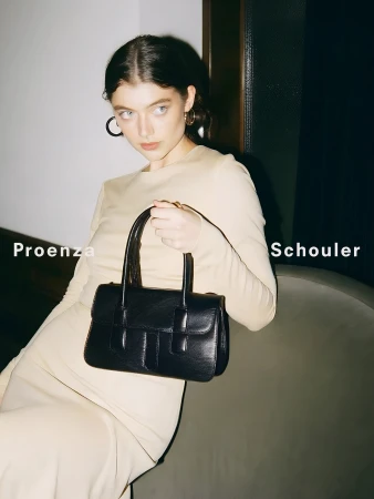 Brunette model holds a black leather handbag in an indoor fashion studio portrait, with visible Proenza Schouler branding on the side.