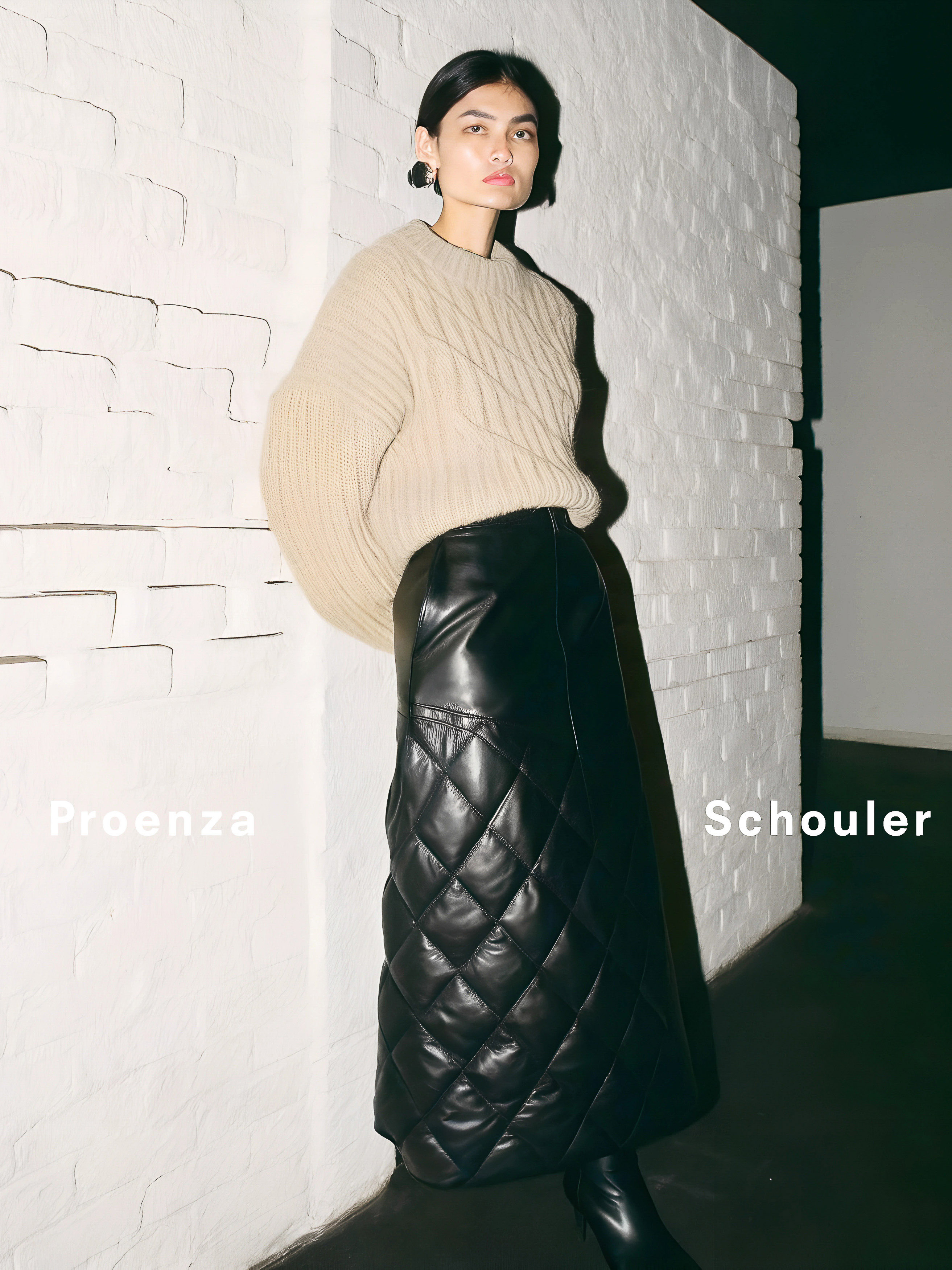 Model wearing a beige knit sweater and long black quilted leather skirt standing against a white painted brick wall.