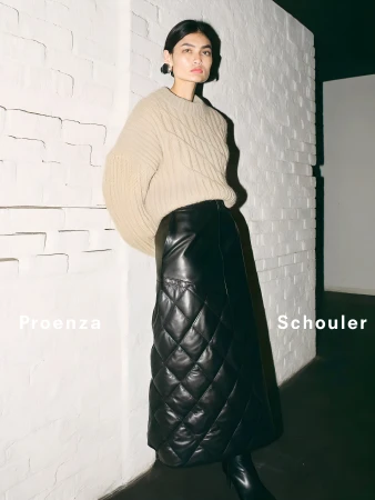 Model wearing a beige knit sweater and long black quilted leather skirt standing against a white painted brick wall.