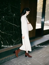 Woman walking outdoors wearing a long cream turtleneck knit dress, brown block-heel ankle boots, and carrying an off-white shoulder bag.