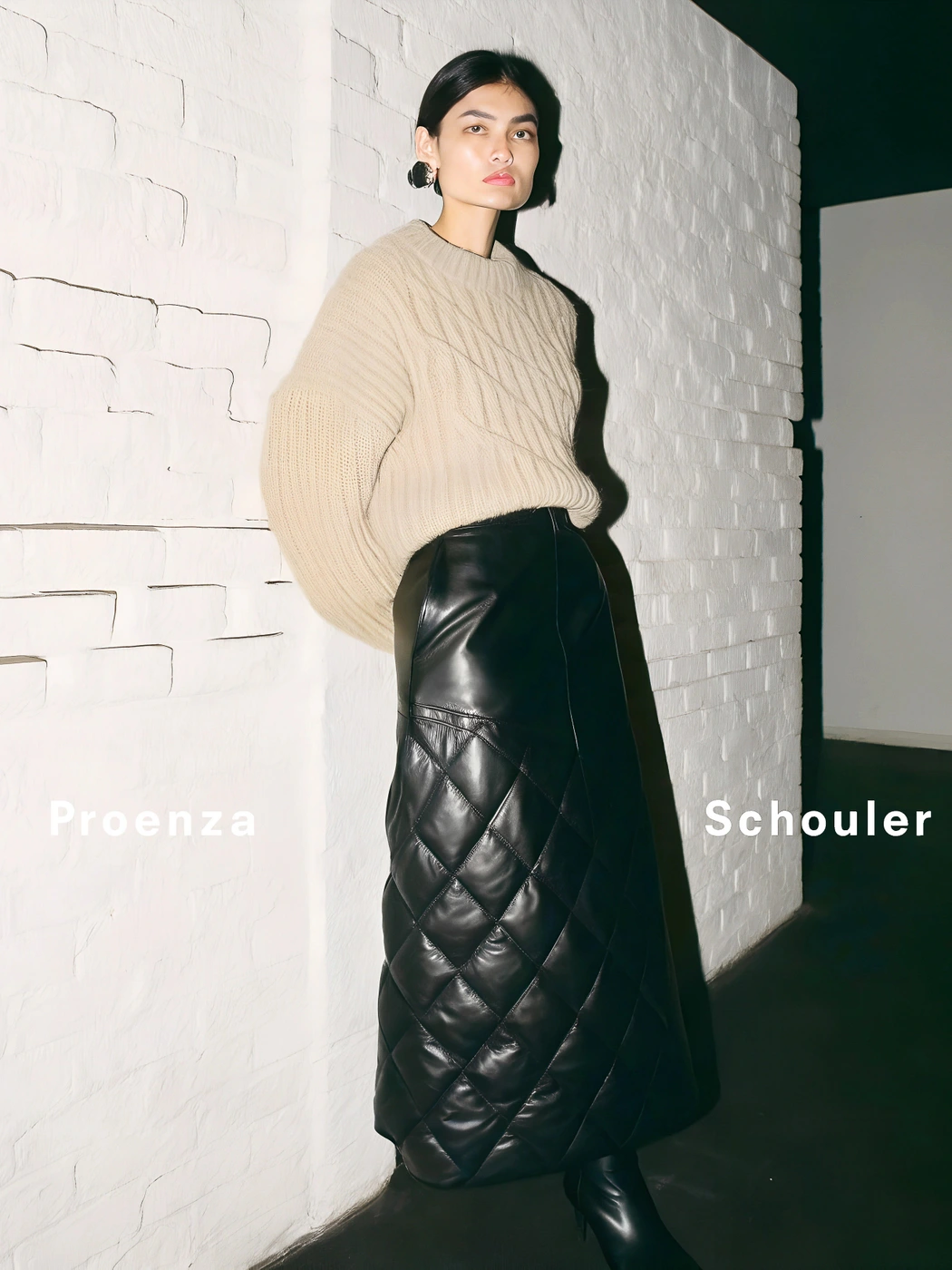 Model wearing a beige knit sweater and long black quilted leather skirt standing against a white painted brick wall.