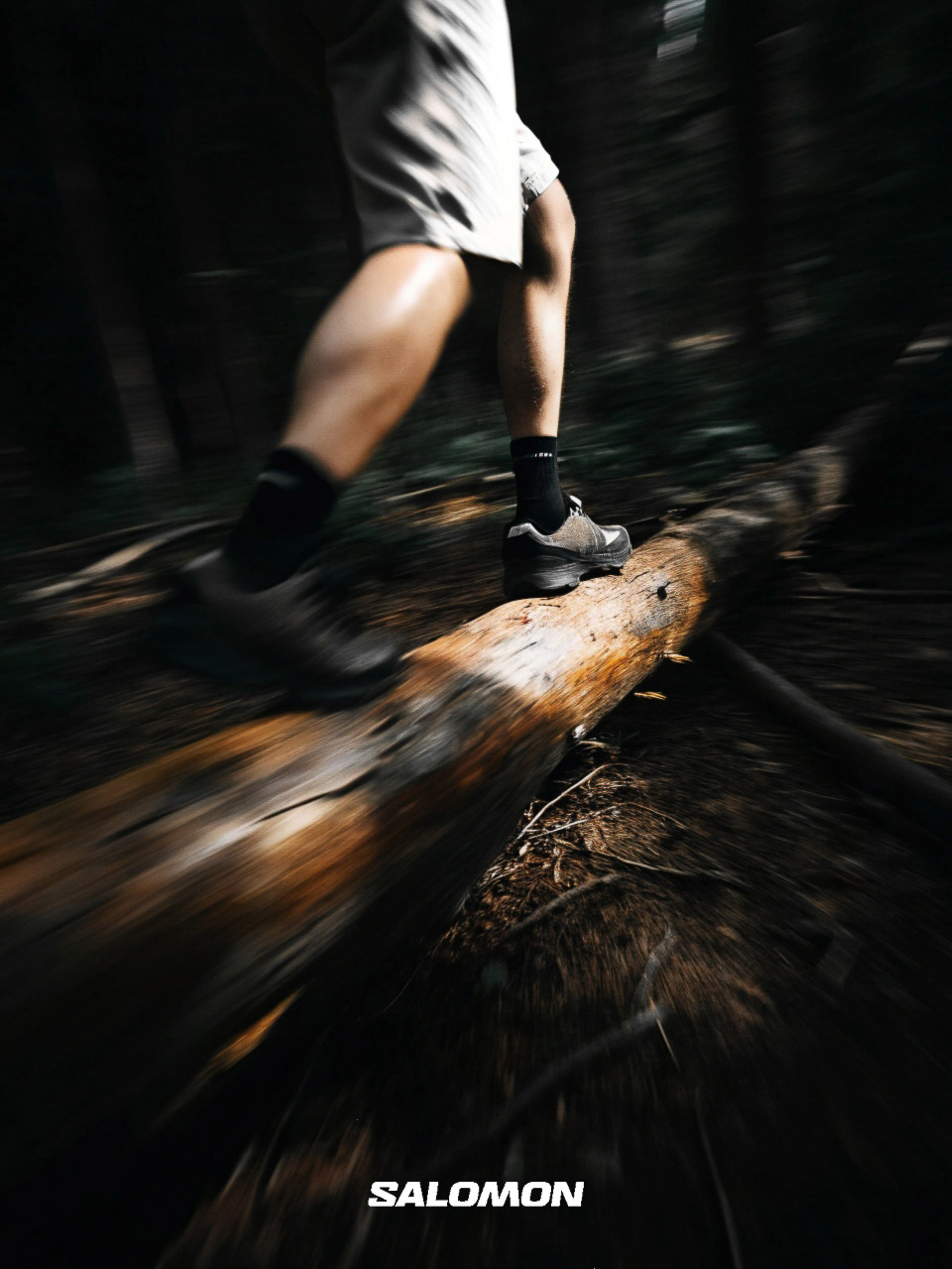 Low-angle motion shot of a man in shorts and athletic shoes walking across a fallen tree trunk in a dark forest setting.