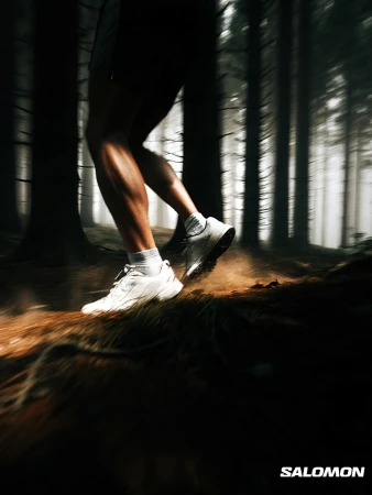 Low angle of a runner's legs and feet in motion on a dirt path surrounded by tall trees in a forest, showcasing athletic footwear.