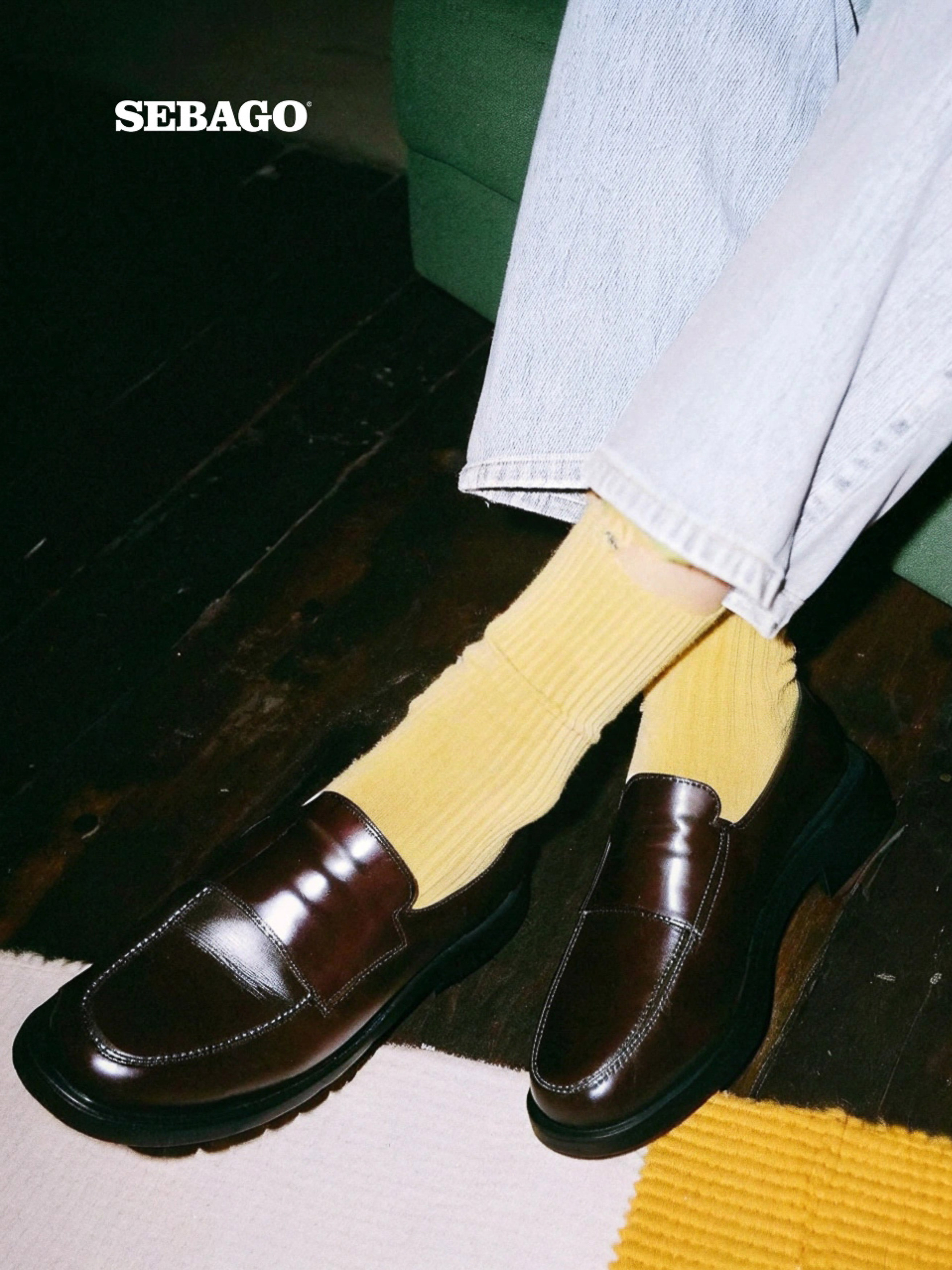 Dark brown leather penny loafers sit beside legs clad in light wash denim jeans and ribbed yellow socks on a dark wood floor.