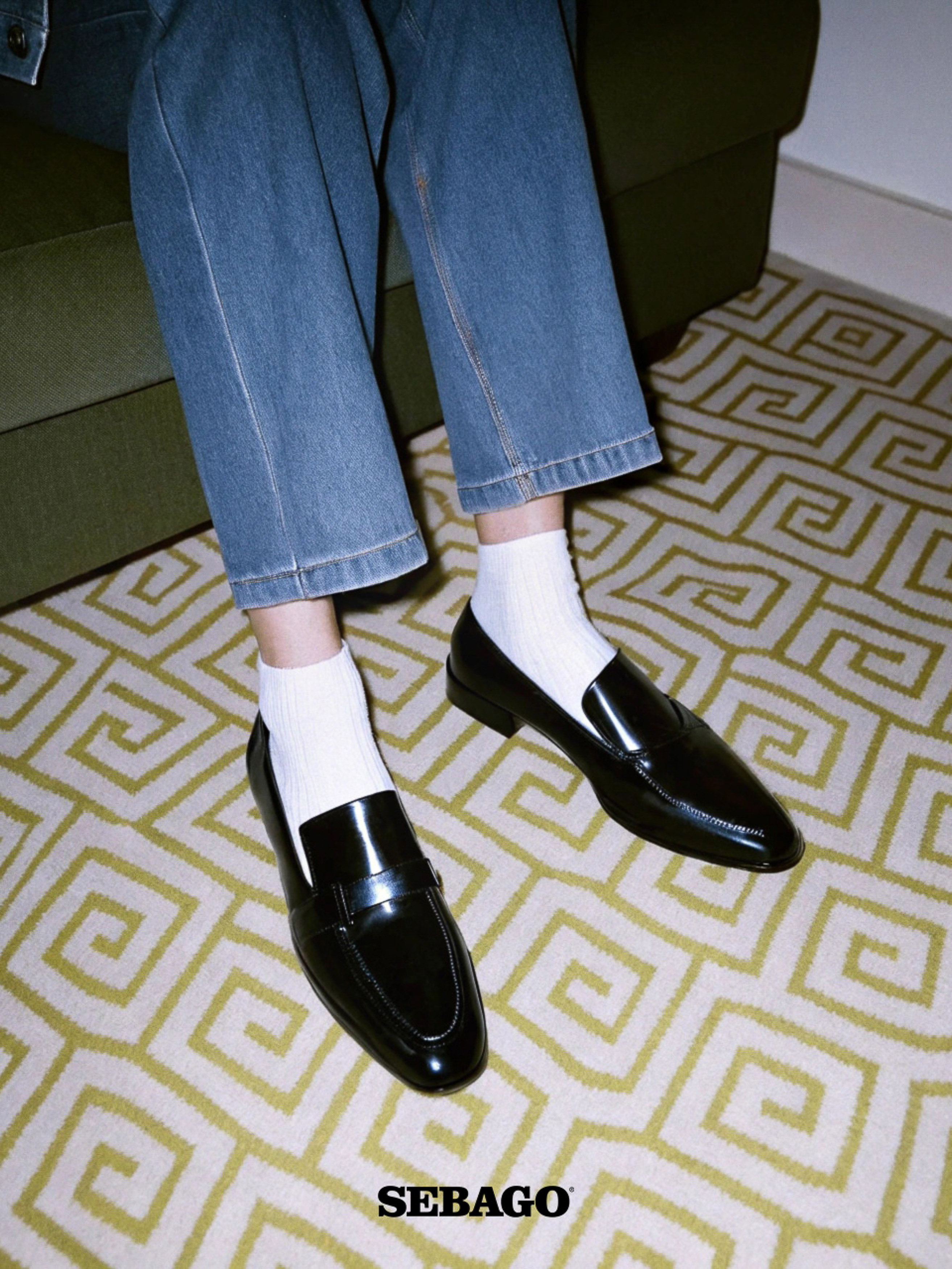 Close-up of black leather loafers paired with white ribbed socks and blue denim jeans against a geometric patterned rug.