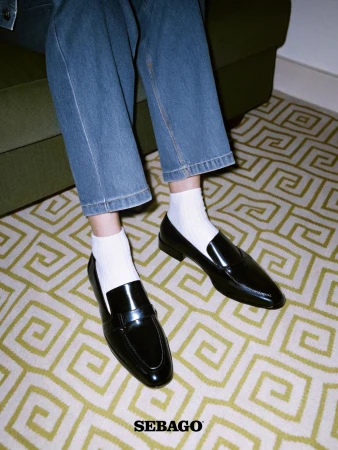 Close-up of black leather loafers paired with white ribbed socks and blue denim jeans against a geometric patterned rug.