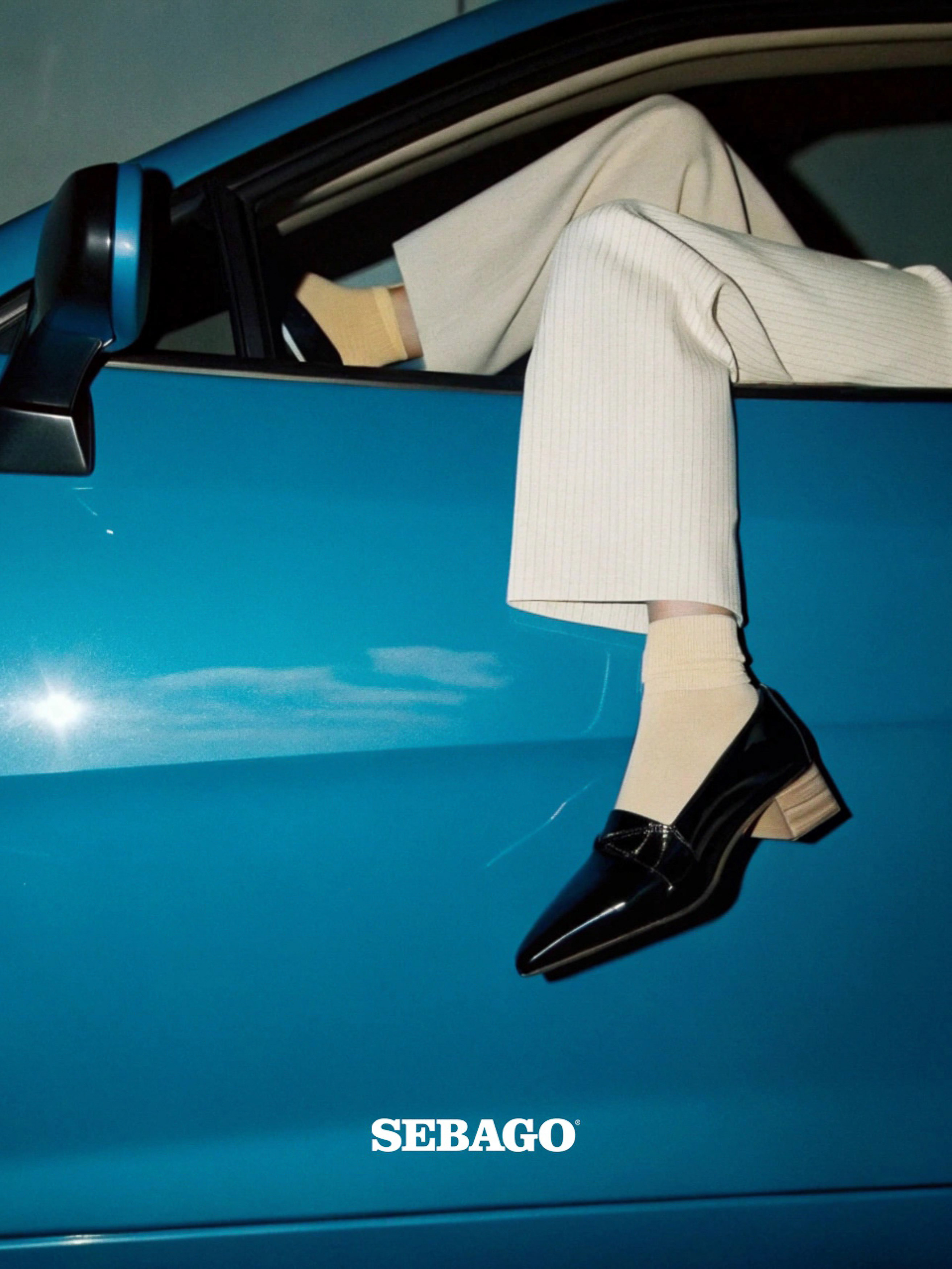 Fashionable shoe ad with a woman's legs and feet dangling from a blue car window, showcasing black loafer shoes and beige socks.