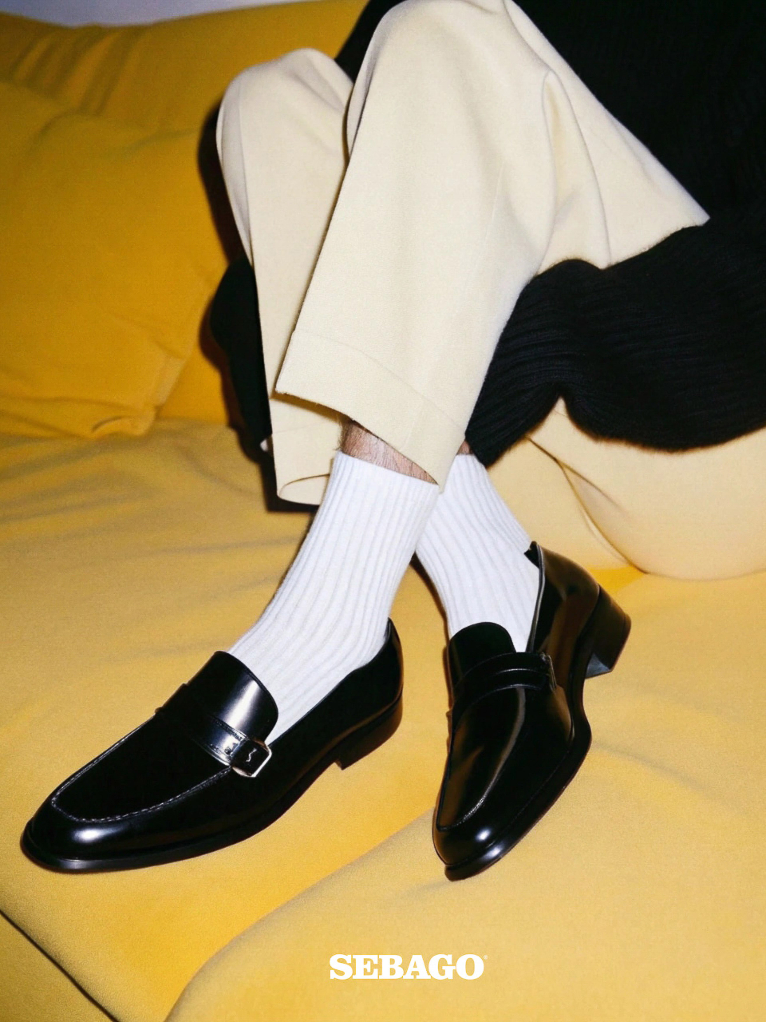 Close-up of two black leather Sebago loafers with white socks on a yellow sofa. Fashion product shot with indoor lighting.