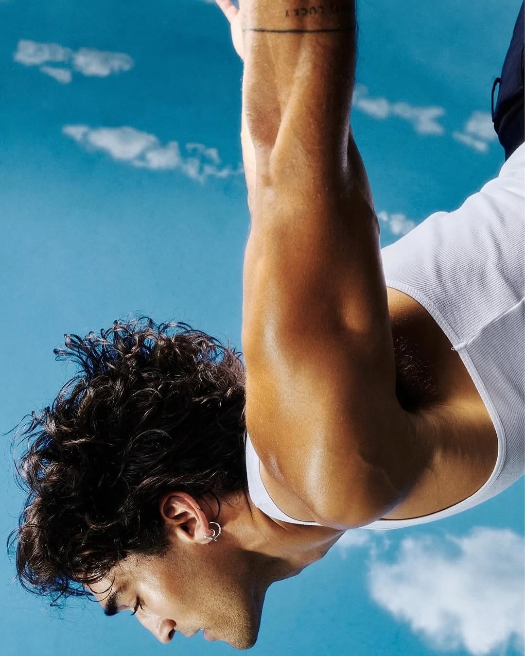 Portrait of a man with curly dark hair in a white tank top against a sky background. Earring visible on ear.